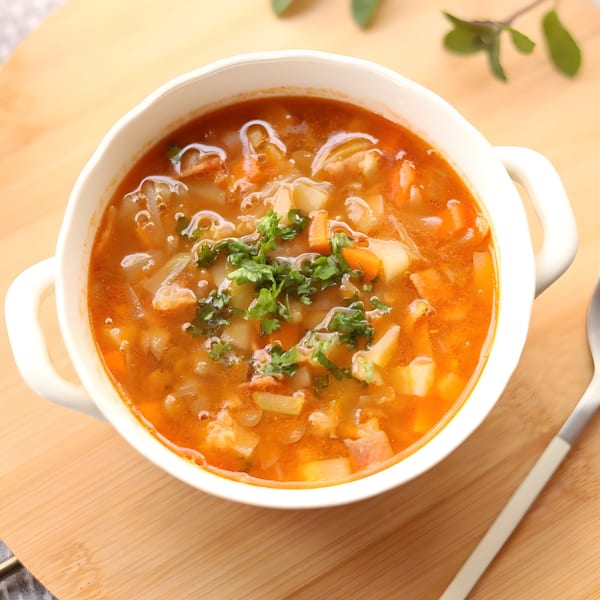 A white bowl of Indian vegetable soup with herbs, carrots, and potatoes on a wooden board.