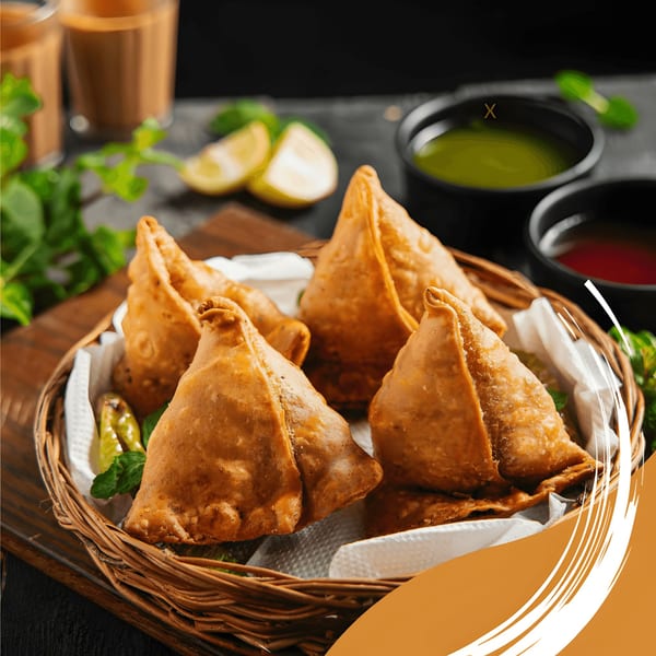 Golden-brown samosas in a woven basket with green and red chutneys, lime, and mint.