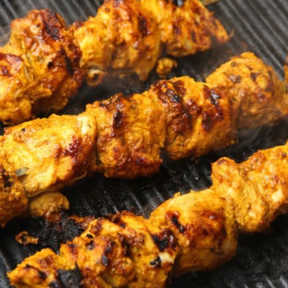 Three skewers of India's Spice chicken tikka grilling on a dark grate, with smoke rising.