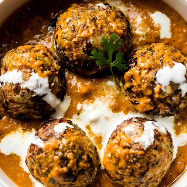 Five Malai Kofta dumplings in a rich orange gravy with white cream swirls and a cilantro garnish.