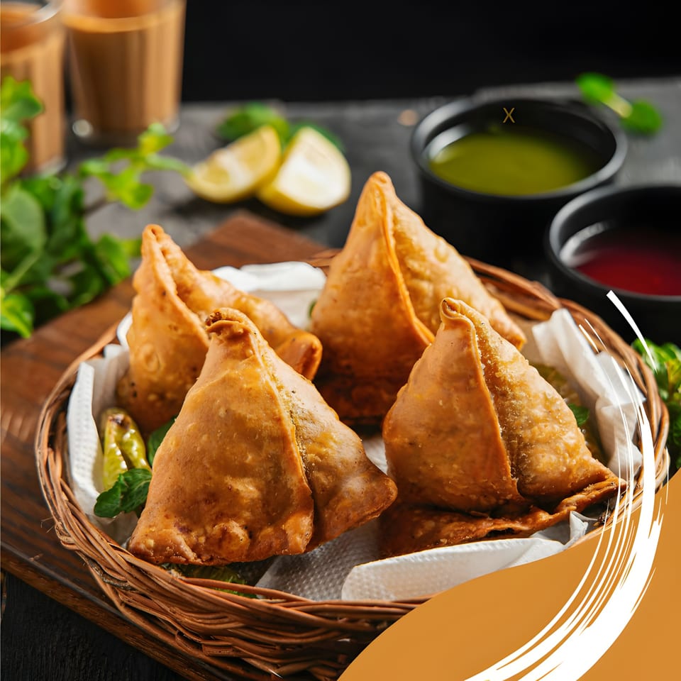 Golden-brown samosas in a woven basket with mint, lime, green chutney, and red chutney.